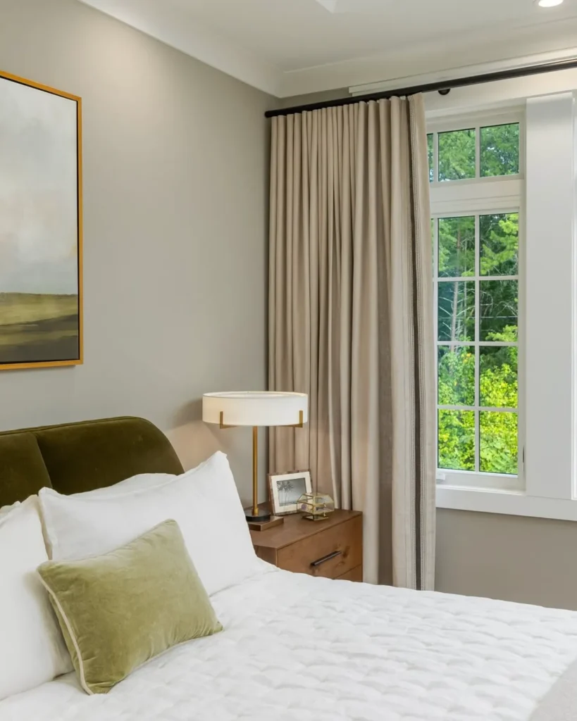 Bedroom with a bed, green pillow, and white bedding. A bedside table holds a lamp and a photo frame. A large window with beige curtains overlooks greenery outside. A painting hangs on the wall.